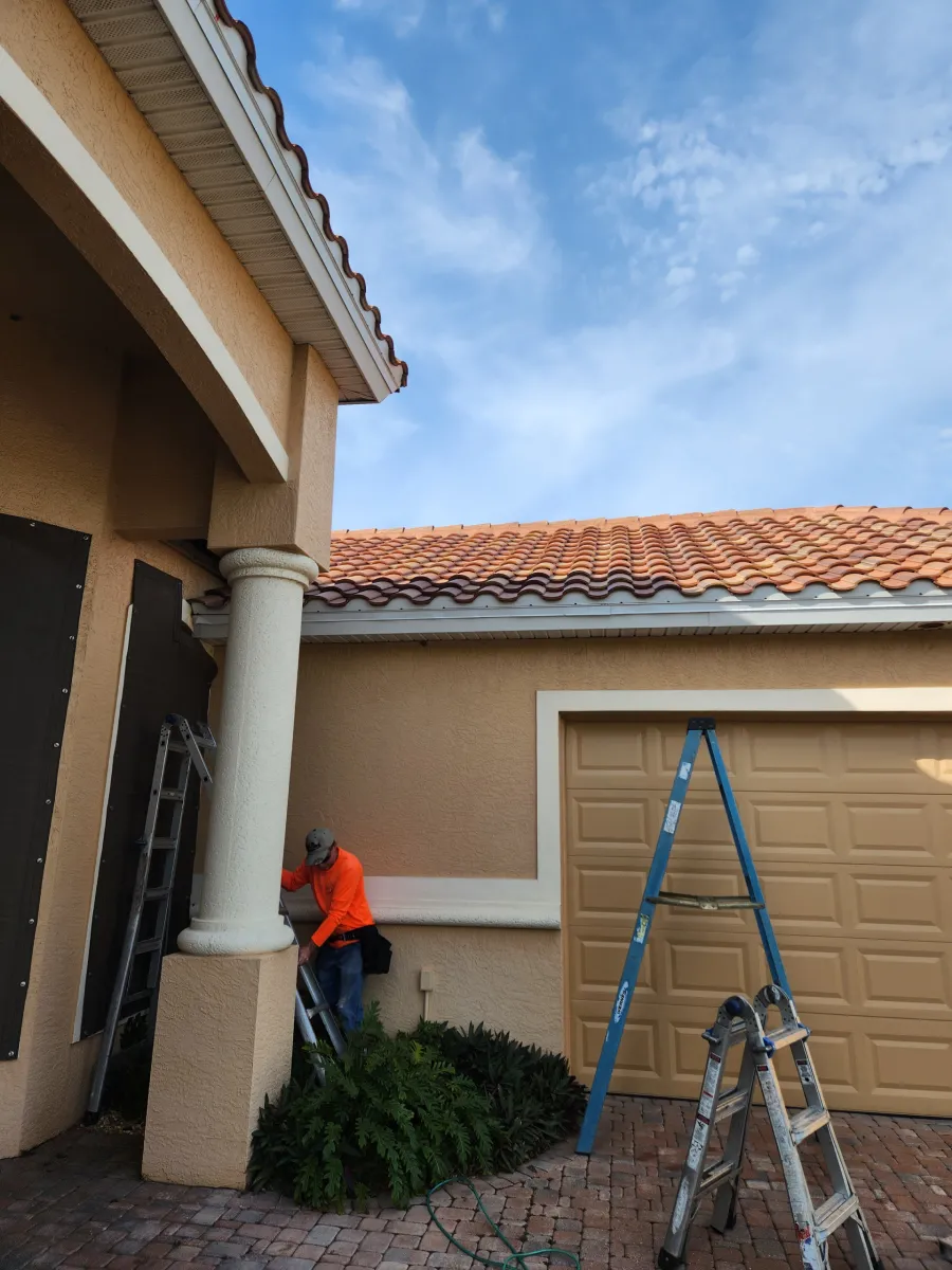 Worker in orange shirt trimming bushes beside a house, with ladder and tools nearby.