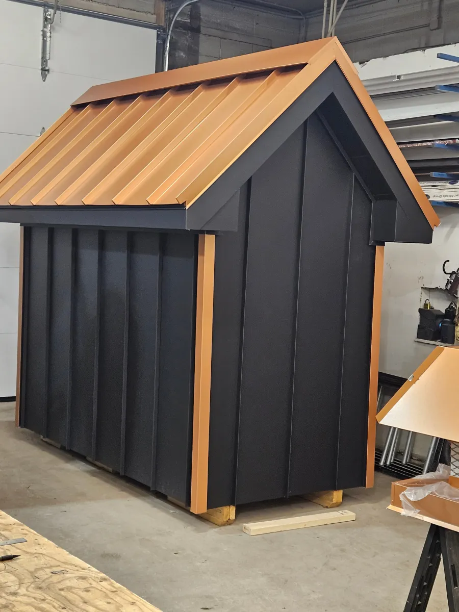 Small black and copper doghouse-style shed in a workshop garage, with corrugated metal siding and roof.