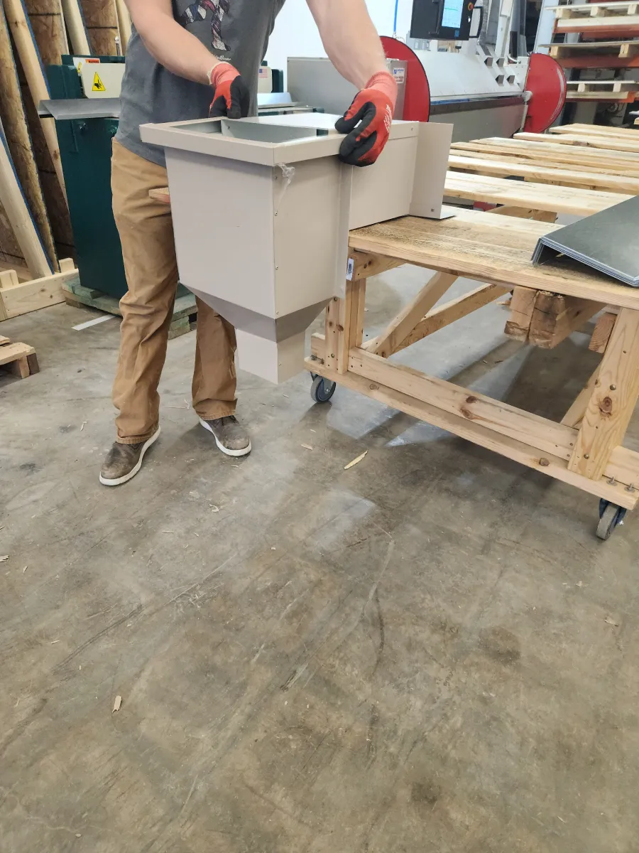 Person holding a gray bin in a workshop beside wooden pallets and a workbench.