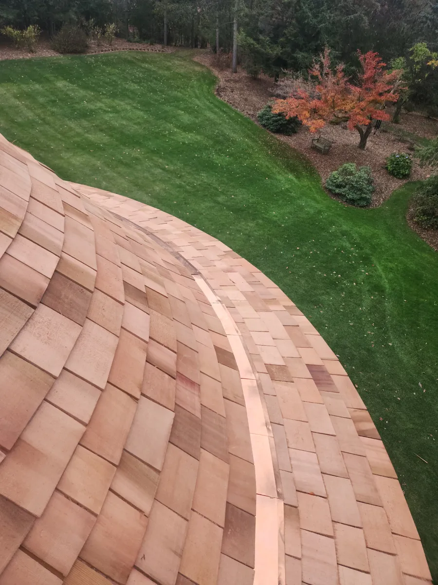 Curved tan roof shingles in foreground, overlooking a green lawn and autumn shrubs