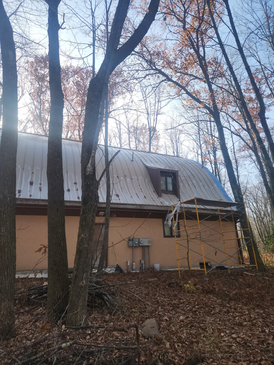 Small brown house with a gray roof among bare trees in a wooded yard.