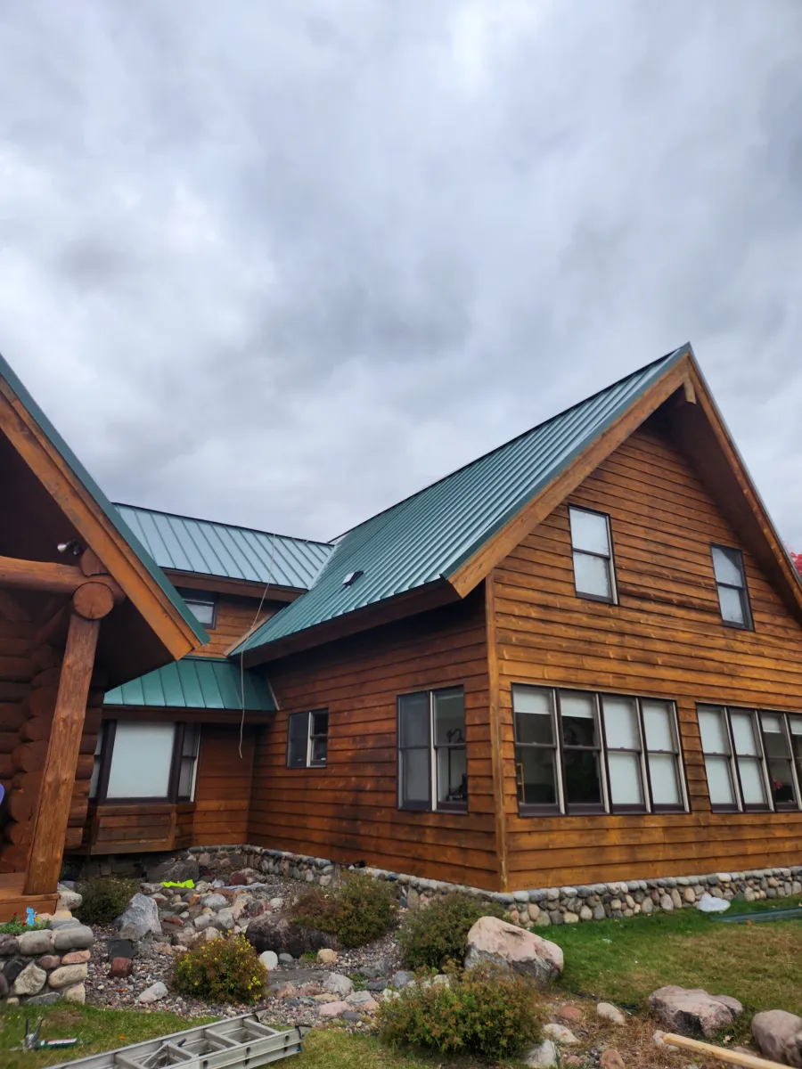 Log cabin with green metal roof and large windows under a cloudy sky, with rocks and grass in front