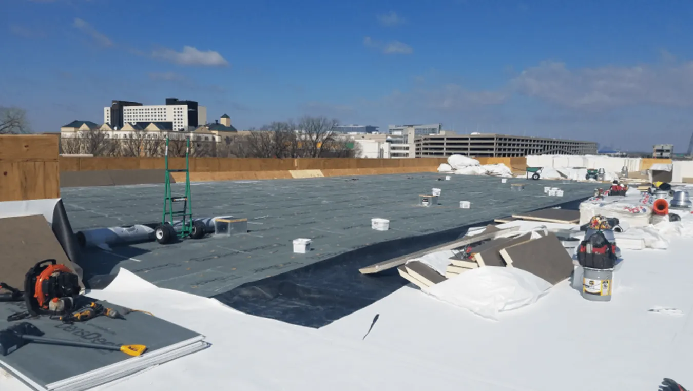 Snow-covered flat rooftop with workers and equipment, black roofing membrane being installed under a clear sky