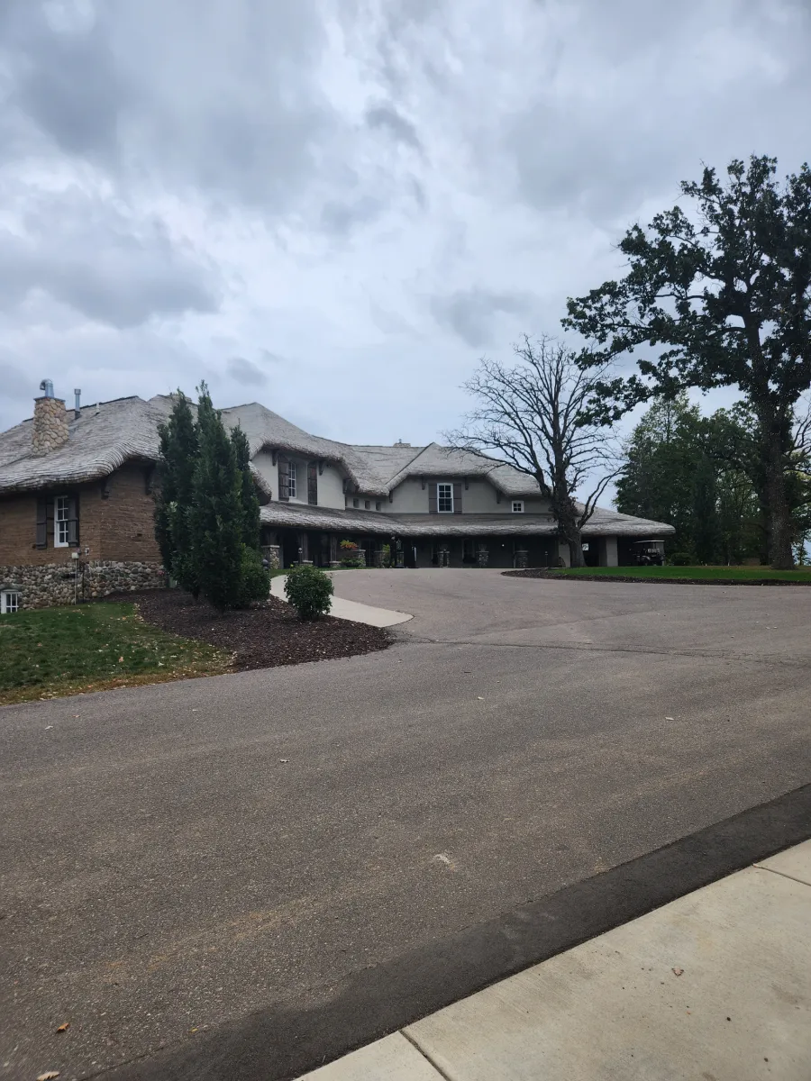 Large stone house with a curved driveway under a cloudy sky, surrounded by trees and landscaping