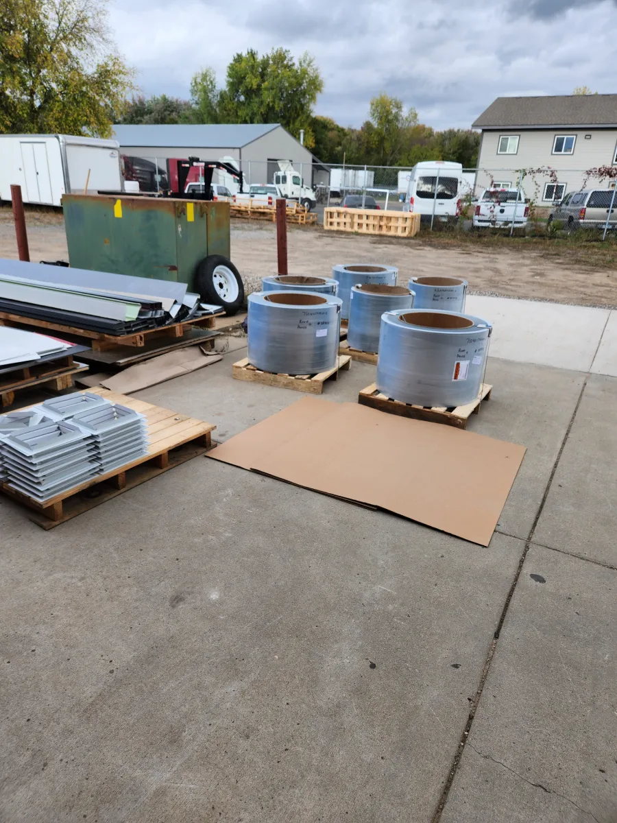 Outdoor industrial yard with metal pipes and three blue barrels on pallets, near a green dumpster and houses.