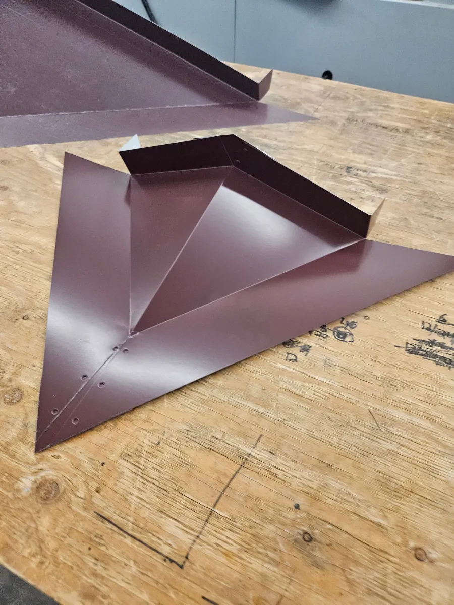 Maroon folded metal sheet sculpture on a wooden workbench, with another folded piece in the background