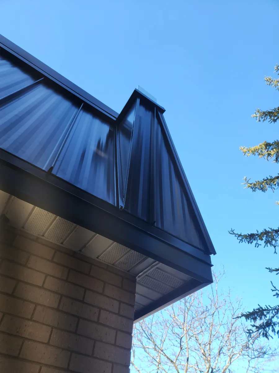 Blue metal roof edge and brick wall of a house against a clear blue sky with bare tree branches