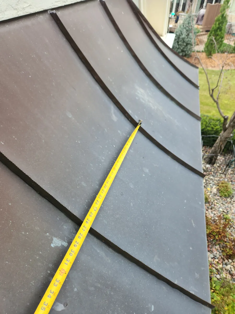 Close-up of a metal roof edge with a yellow measuring tape and dark curved panels.