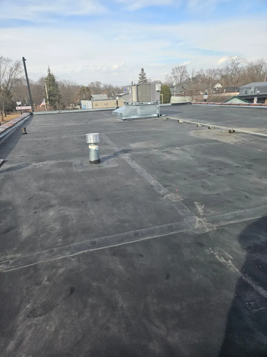 Flat rooftop with black membrane, vents, and distant trees under a cloudy sky.