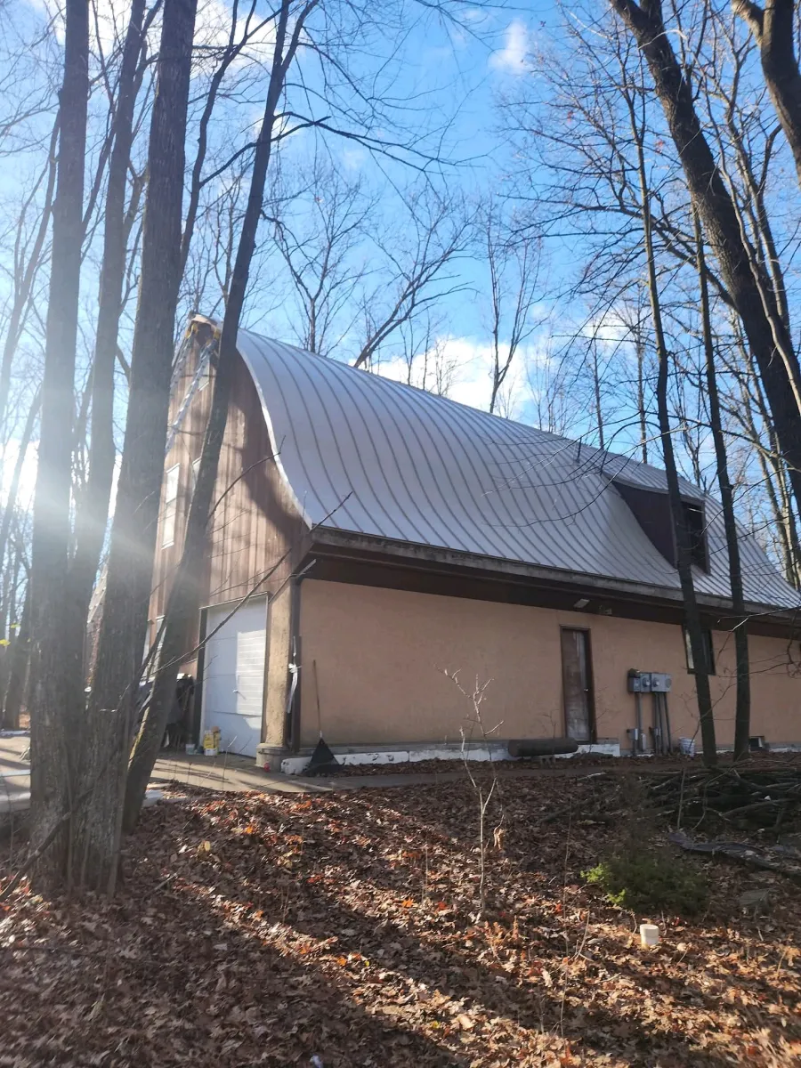 A beige cabin with a metal roof in a leaf-covered wooded area under a bright blue sky.