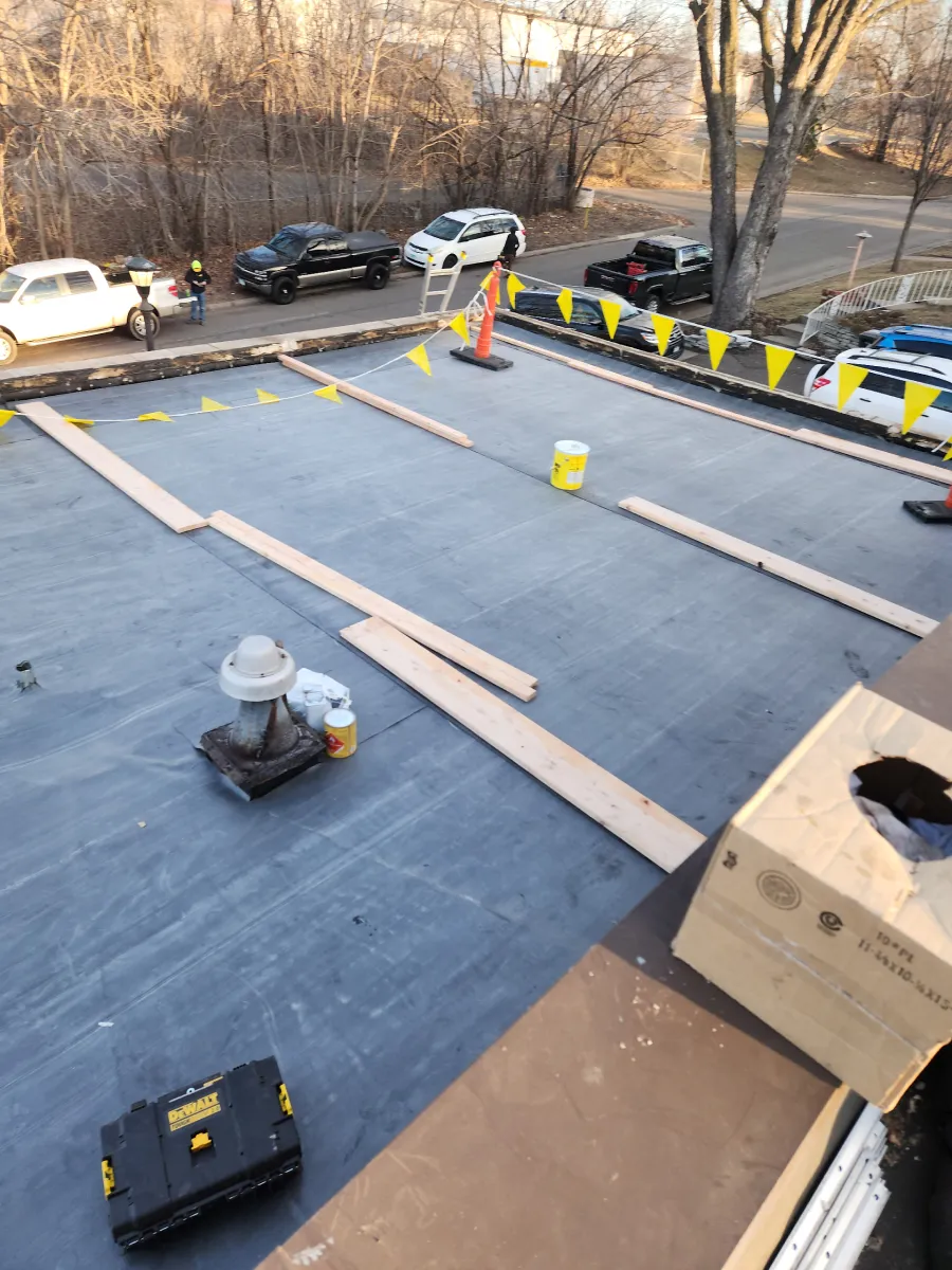 Flat rooftop under construction with black membrane, lumber strips, tools, and parked cars below.
