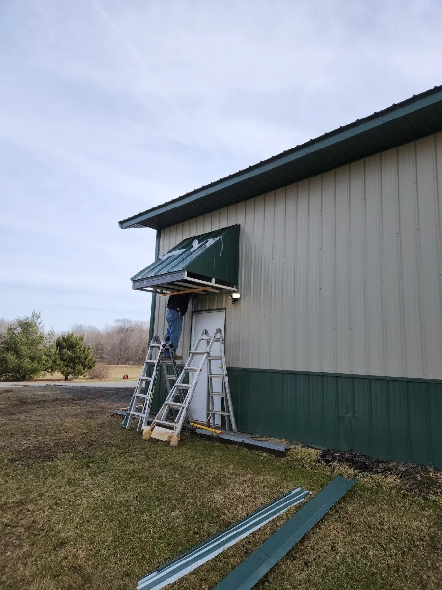 Workers on ladders repairing a green awning on a metal building exterior.