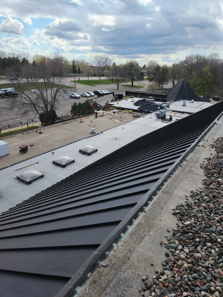 Rooftop under construction with black metal panels, gravel edges, and workers in the distance under a cloudy sky