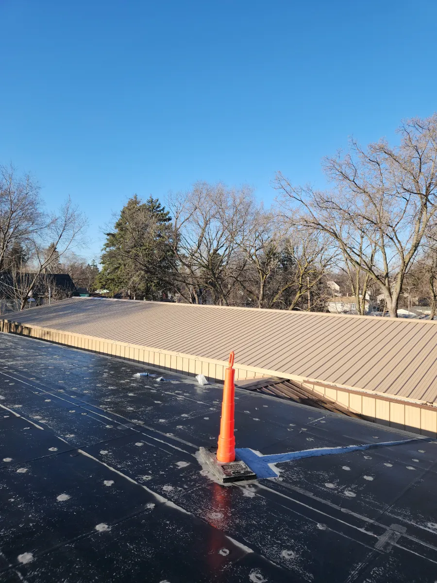 Orange traffic cone on a black rooftop with a metal vent, trees and blue sky in the background