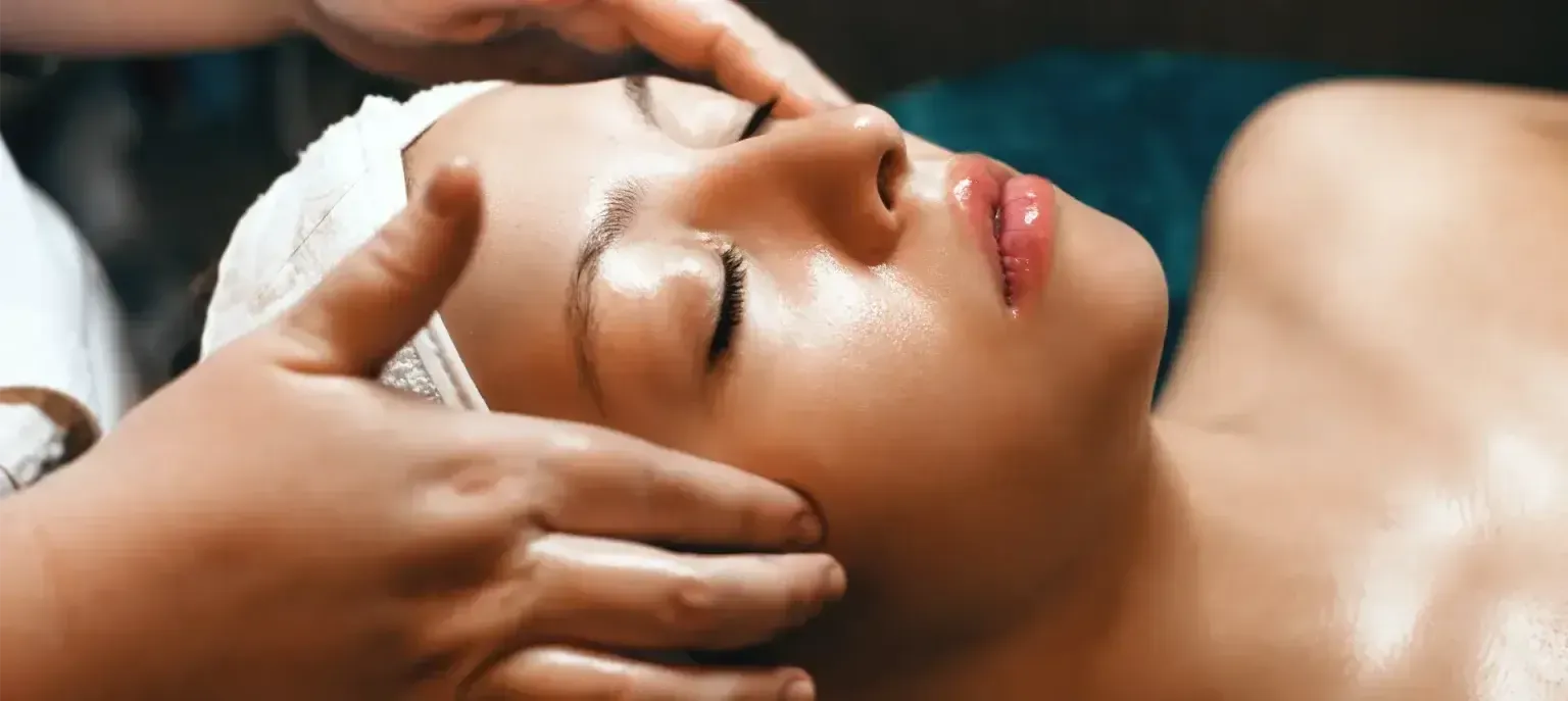 Person receiving a facial massage with closed eyes, wrapped in a white towel and foam headband