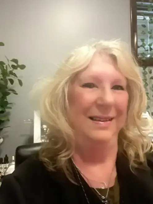 Smiling woman with blonde hair in an indoor office setting, wearing a black top and necklace.