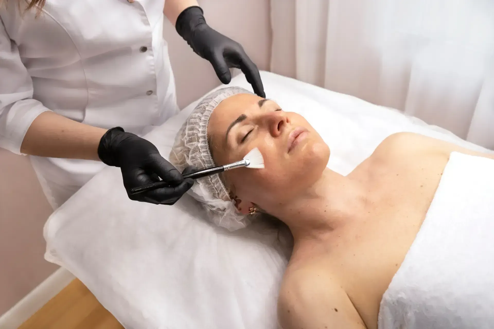 Esthetician applying facial treatment to client lying on spa bed, wearing black gloves and headband.
