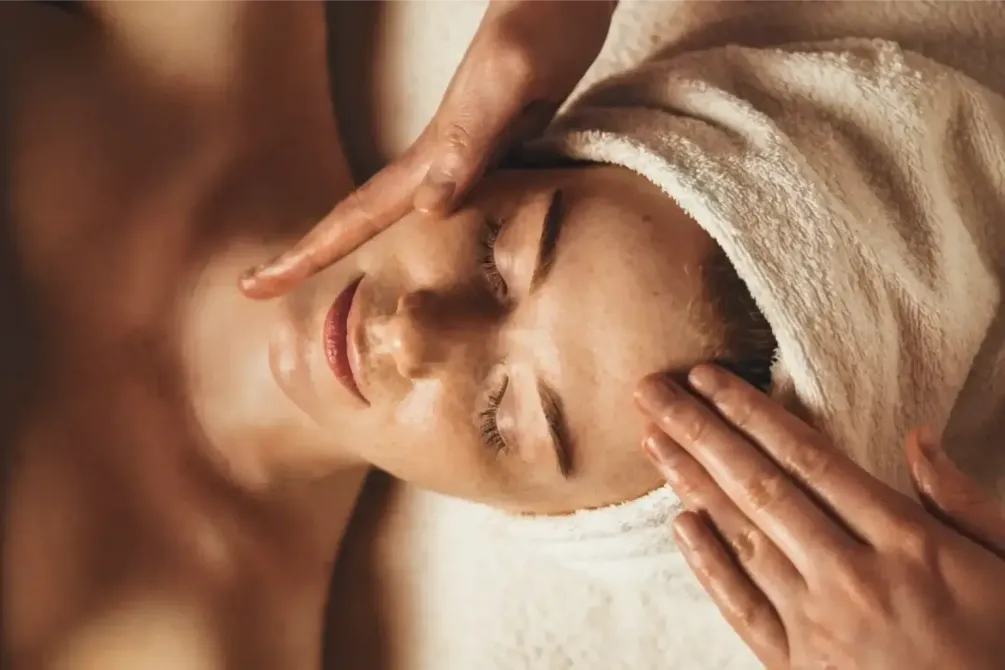 Facial spa treatment with a therapist massaging a client’s forehead under a white towel wrap