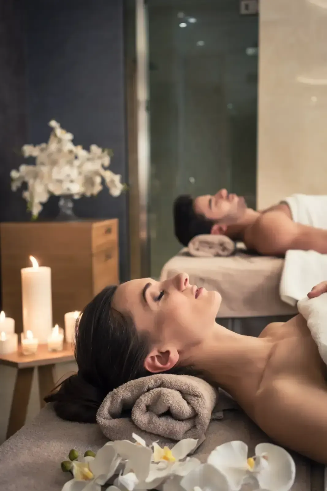 Two people relaxing on massage tables in a candlelit spa with white flowers and towels