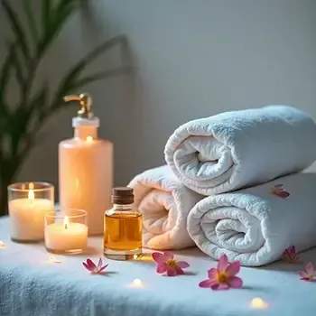 Spa setup with rolled white towels, candles, oil bottle, and pink flowers on a soft blue surface