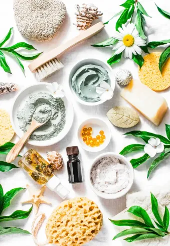 A flat lay of natural skincare products, white bowls, loofah sponges, green leaves, and a white flower.