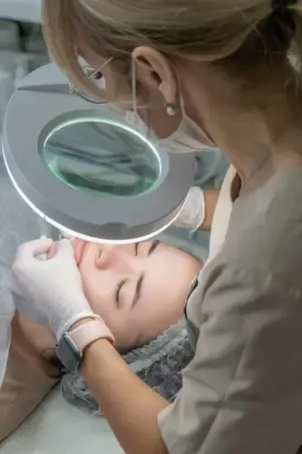Beauty technician inspecting a client’s eyebrow with a magnifying lamp and gloved hands