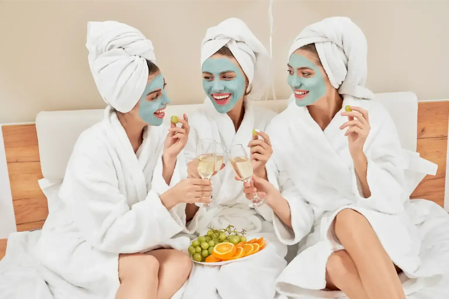 Three friends in white robes and towels wear face masks, holding fruit in a spa-like setting