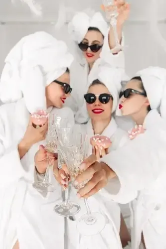 Four women in white robes and towels with sunglasses toast champagne in a bright spa setting