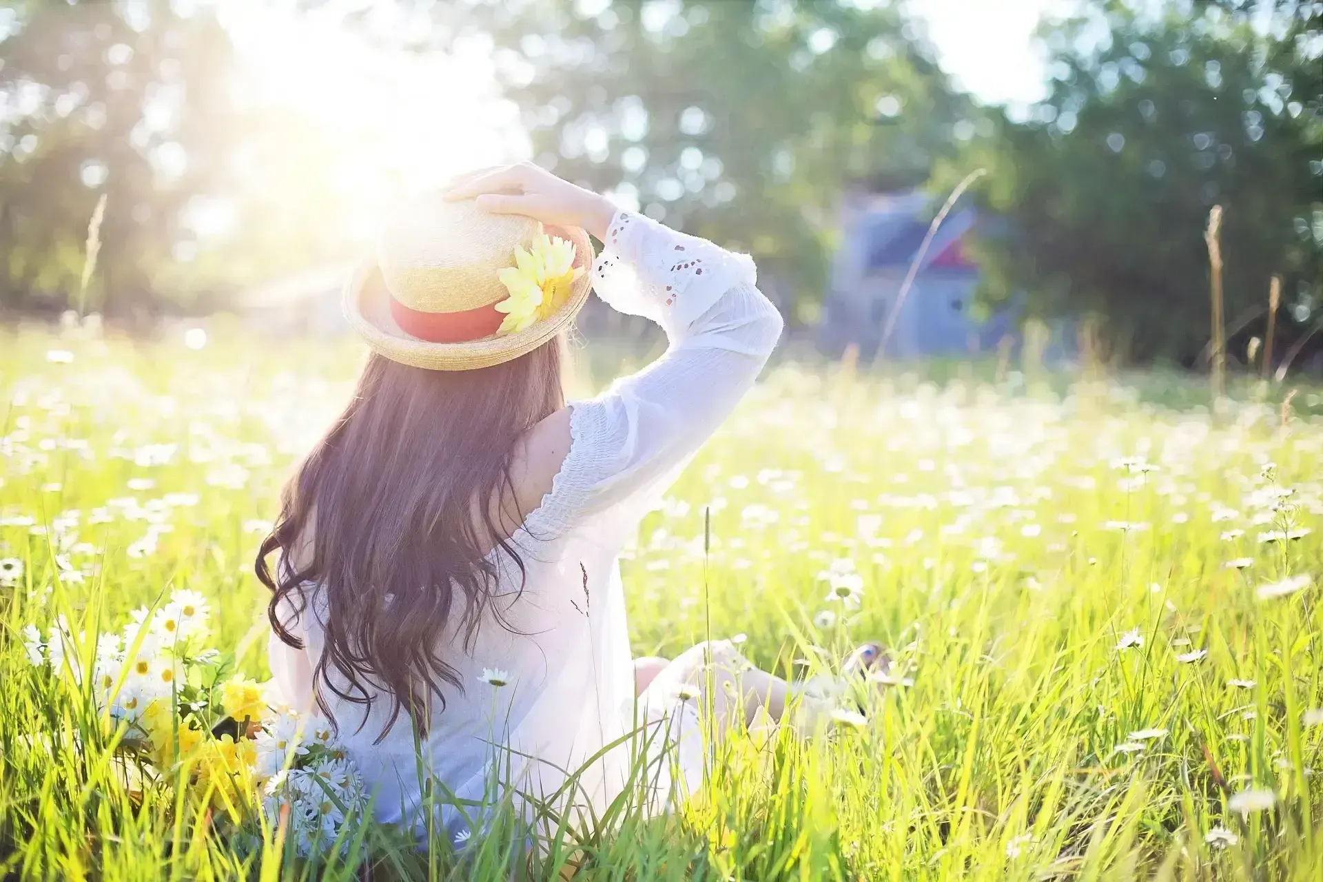 Person sitting in a sunlit grassy field, wearing a straw hat and white shirt, with long hair down back