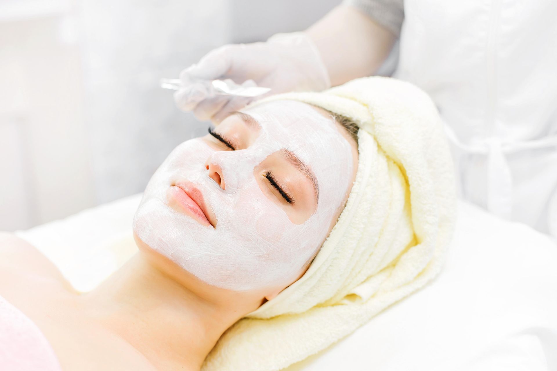 Woman receiving a white facial mask treatment in a spa, wrapped in a cream towel.