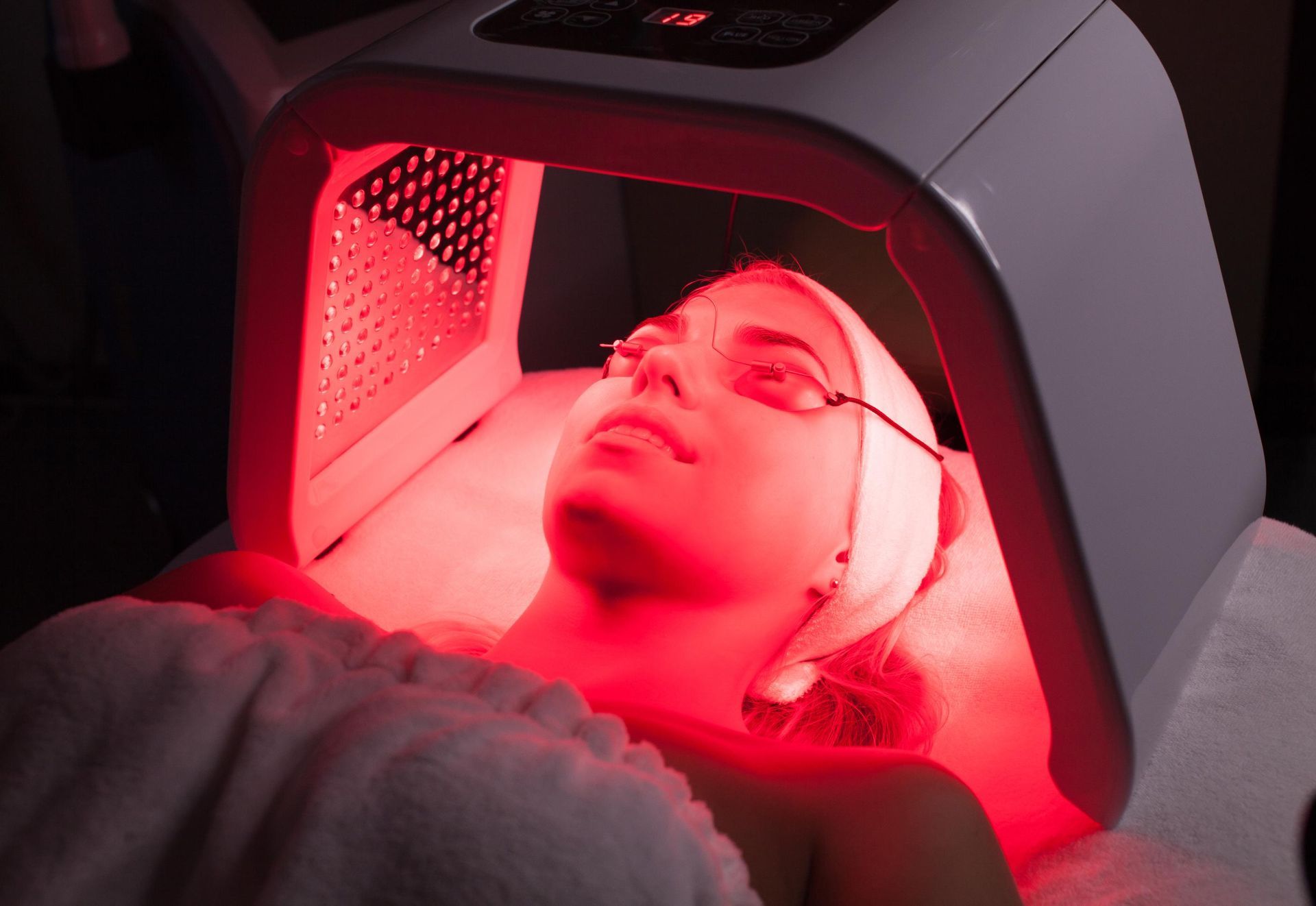 Person lying under a red light therapy device with a face mask on, eyes closed
