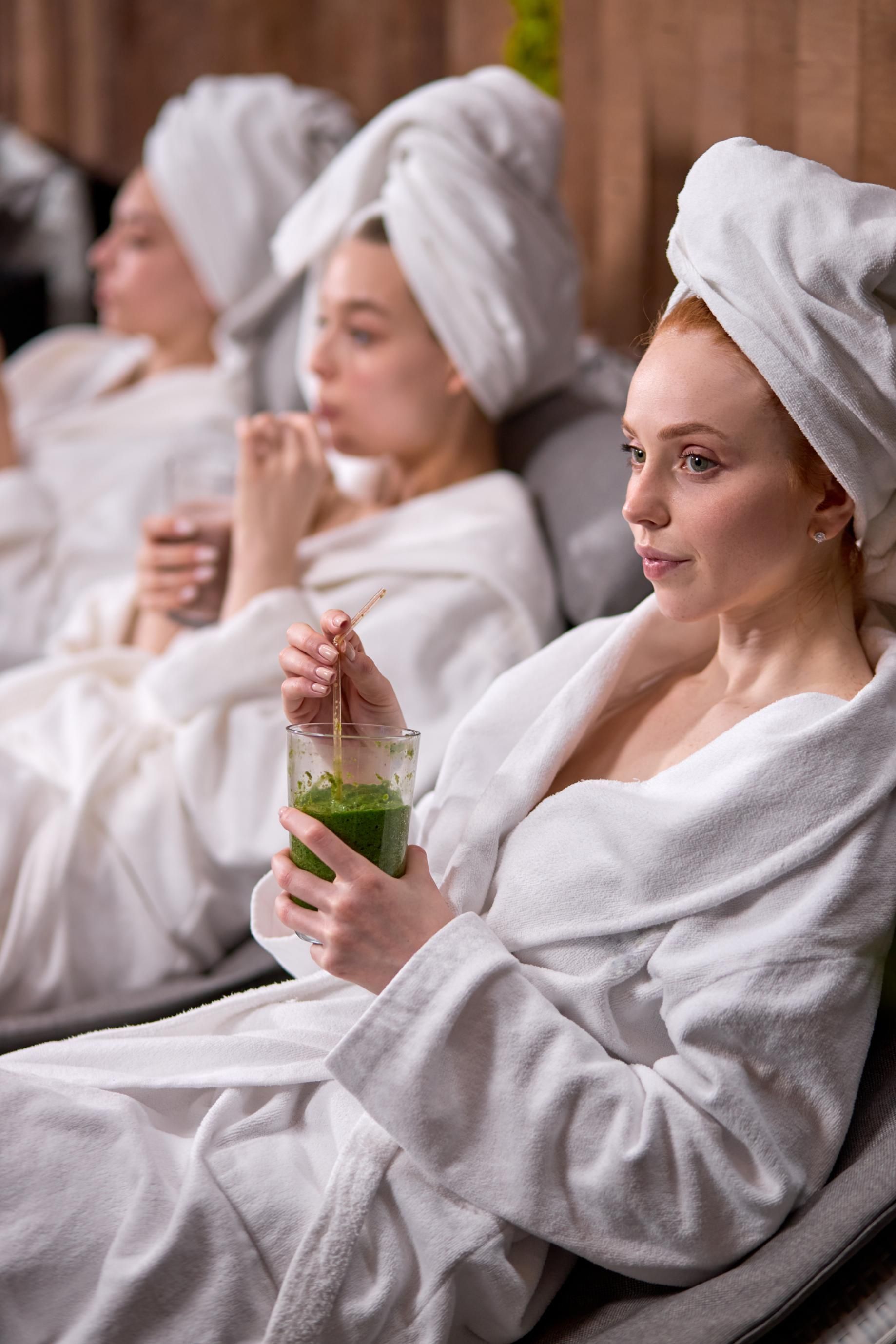 People wrapped in white towels relaxing on benches, holding drinks in a spa-like setting