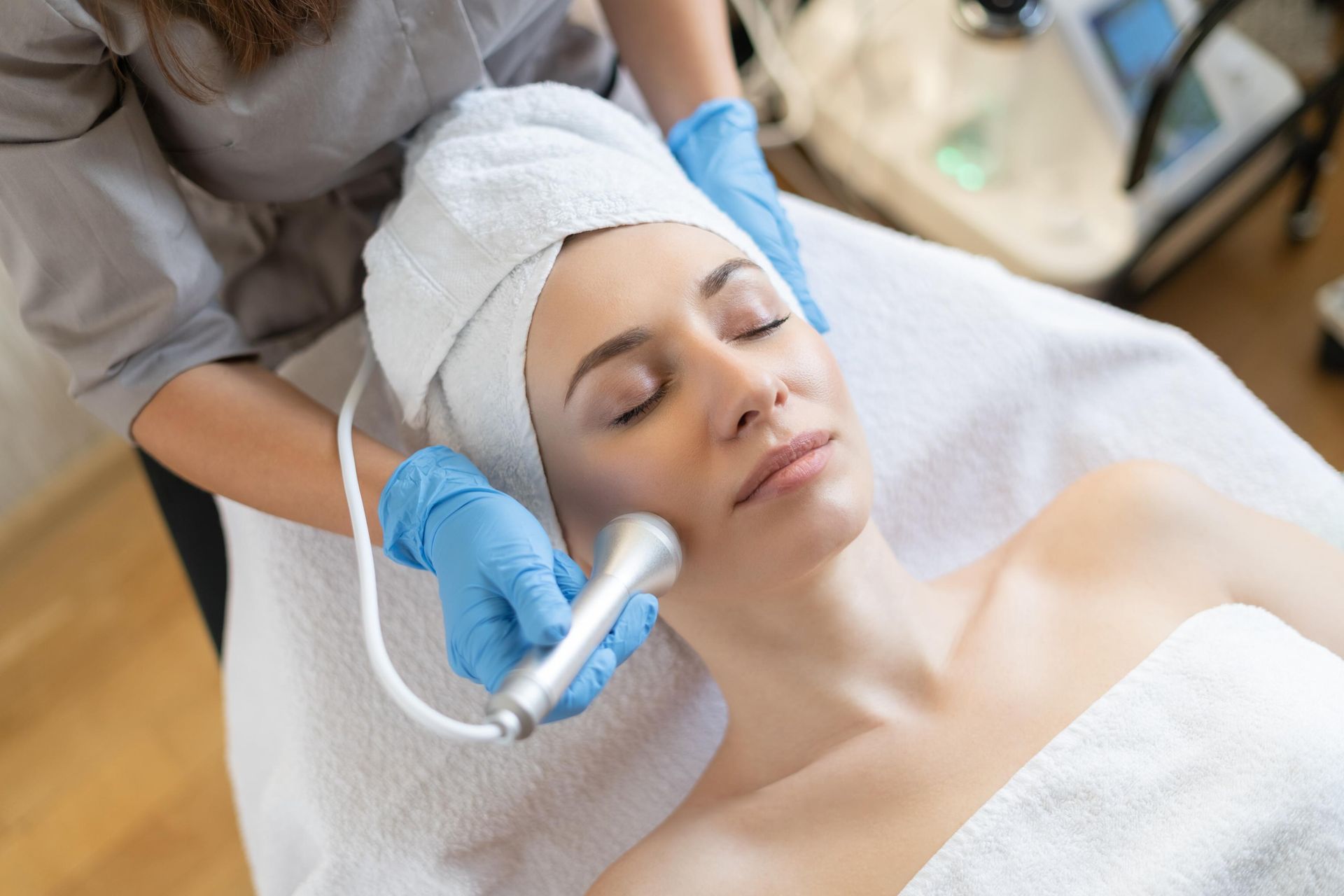 Aesthetic treatment being applied to a woman’s face with a handheld device, wearing a white towel head wrap and blue gloves
