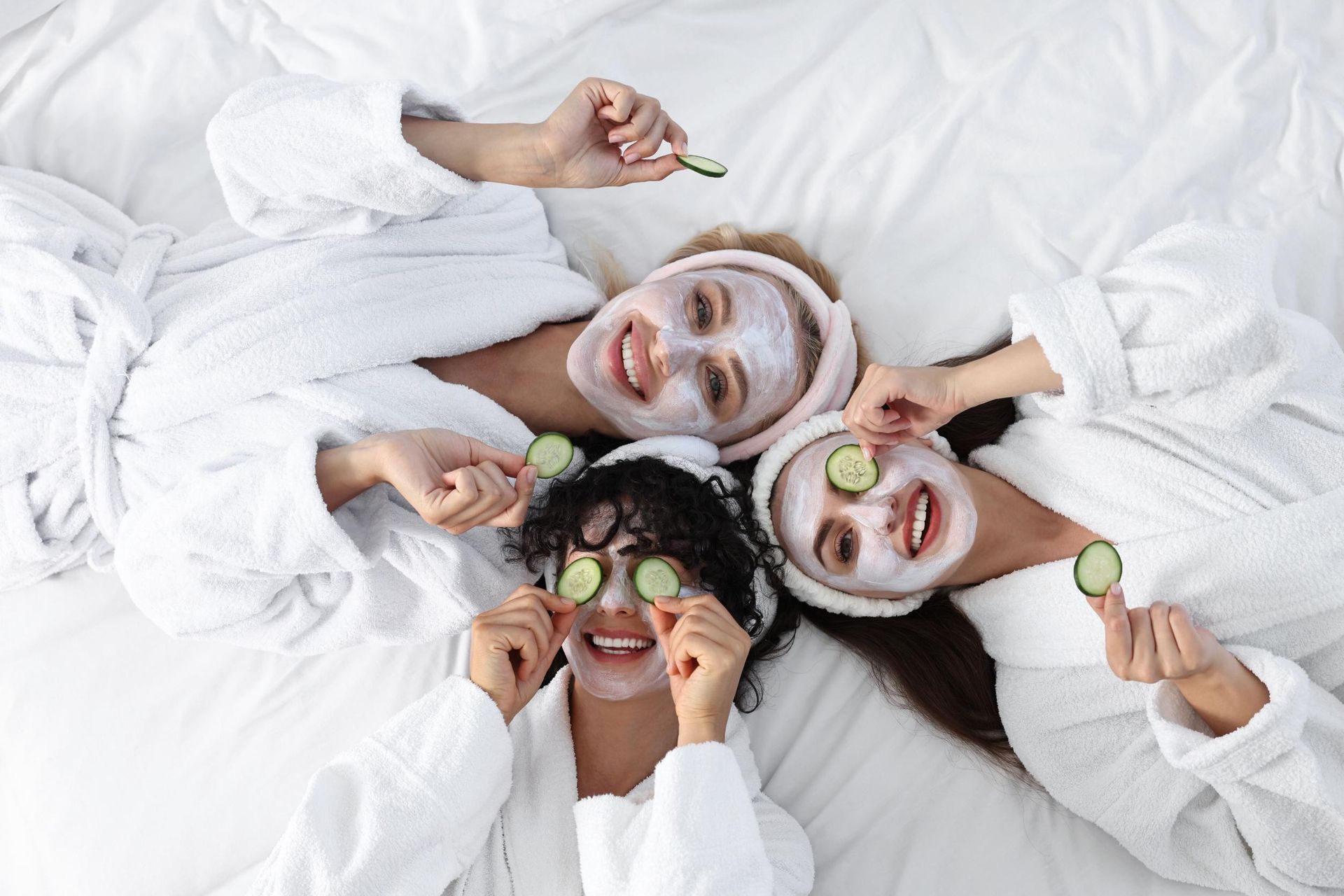 Three friends in white robes lie on a bed with cucumber slices and facial masks, smiling and relaxing