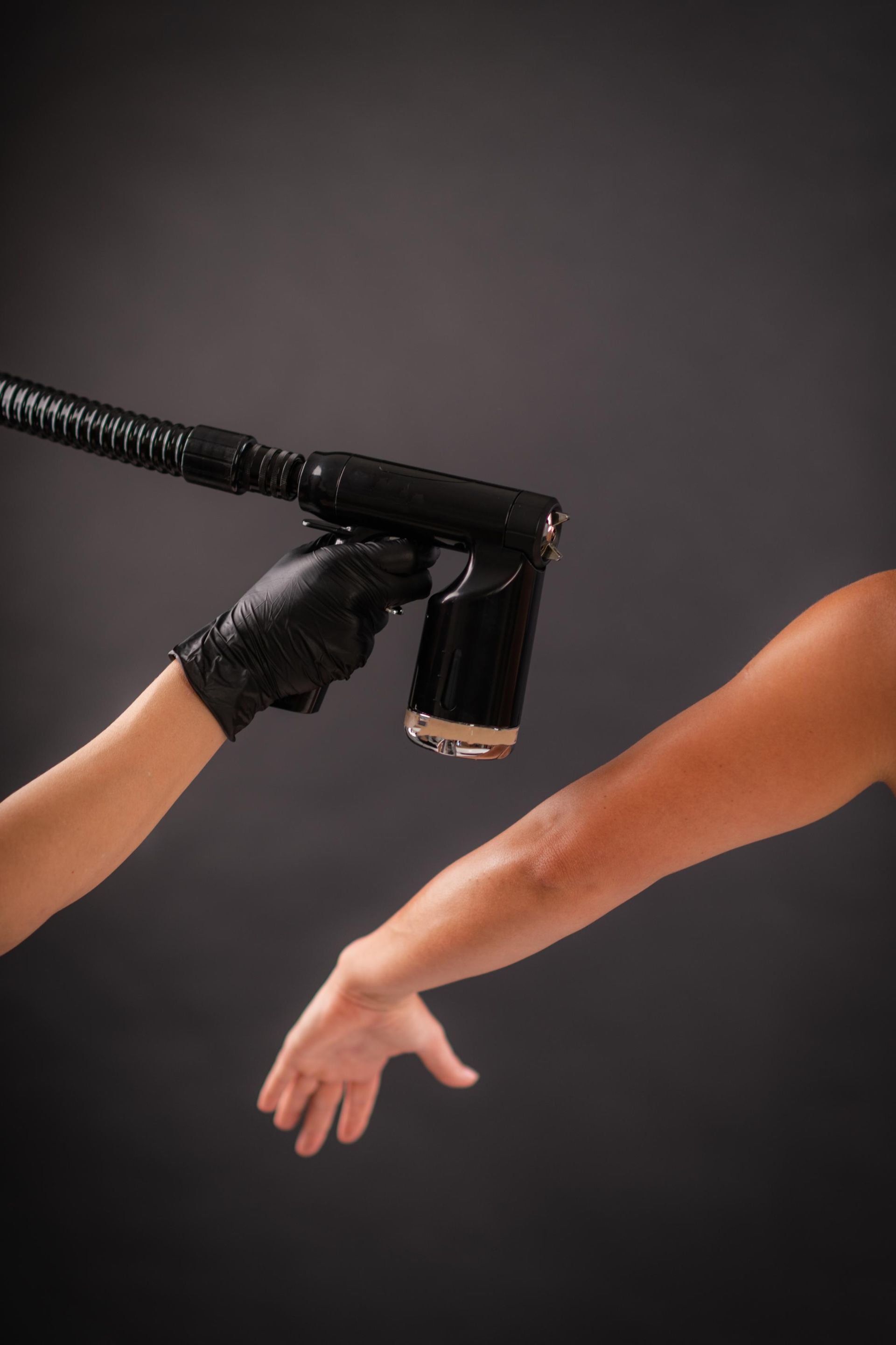 Black spray tanning nozzle coating a person's outstretched arm against a dark background