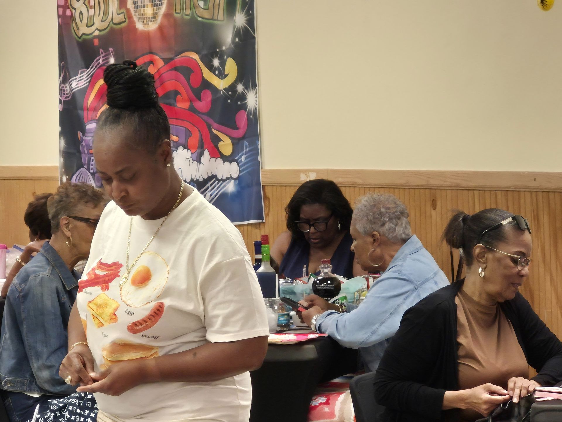 Woman in a white shirt with a food design stands near a table with other people in a decorated room.