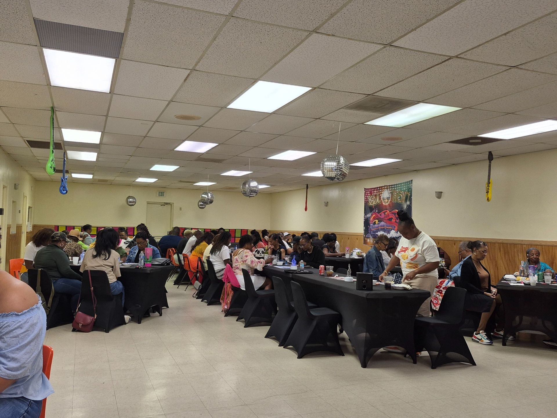 People seated at tables in a brightly lit room. Some are eating or interacting, decorations hang from the ceiling.