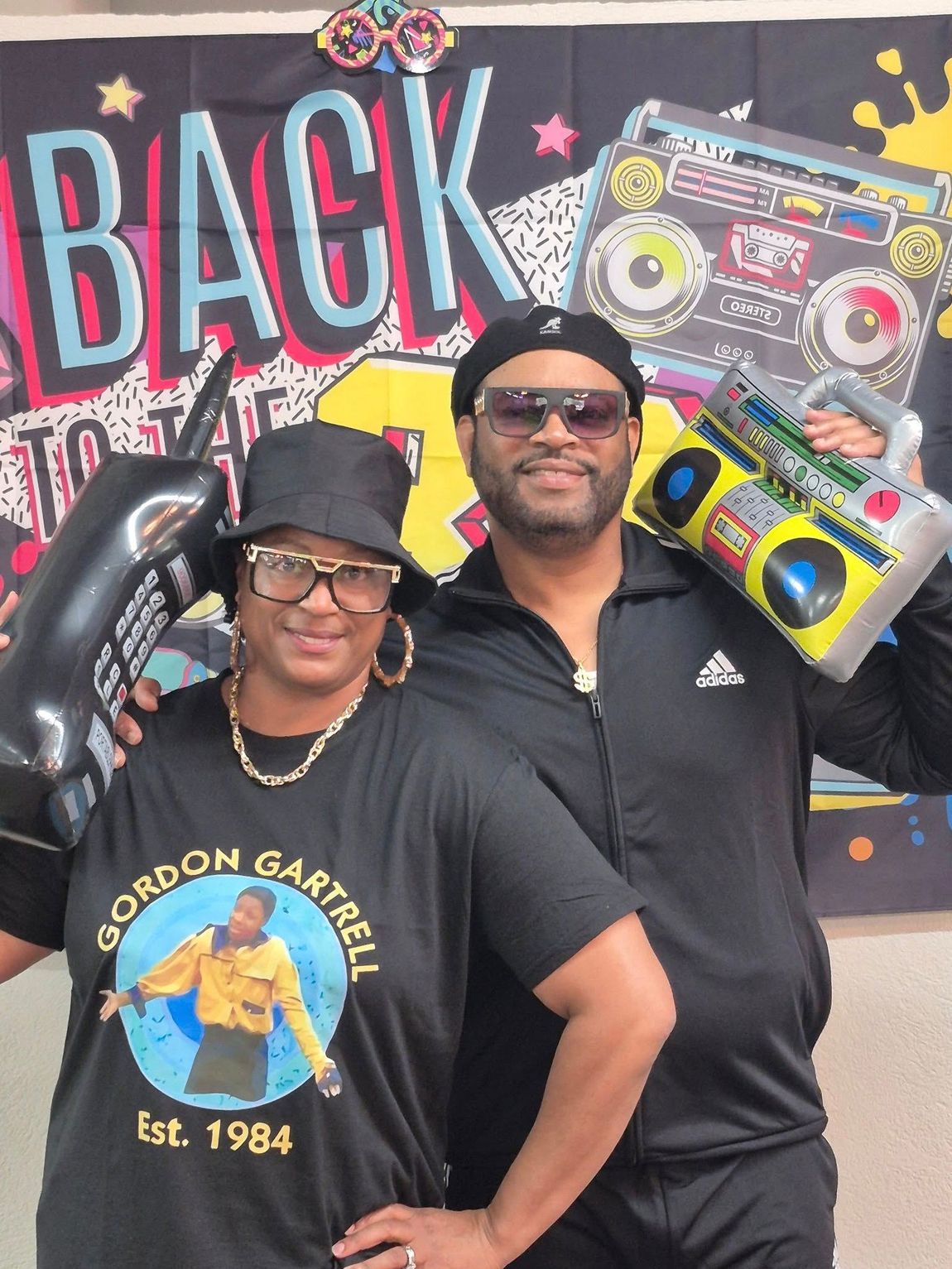 Two people in 80s attire, posing in front of a Back to the Future backdrop with props like a boombox and inflatable phone.