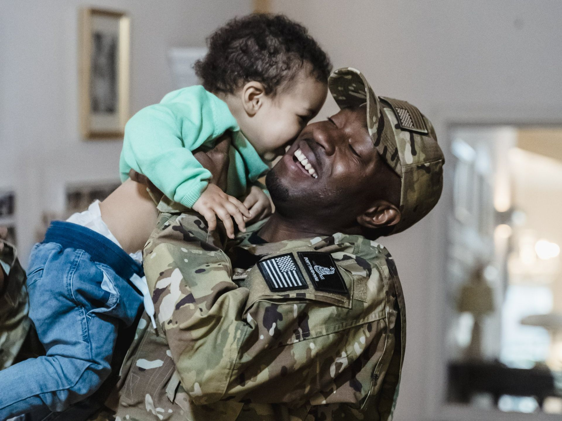 Soldier in uniform smiles while holding a baby, both touching faces indoors in Spring Lake, NC.