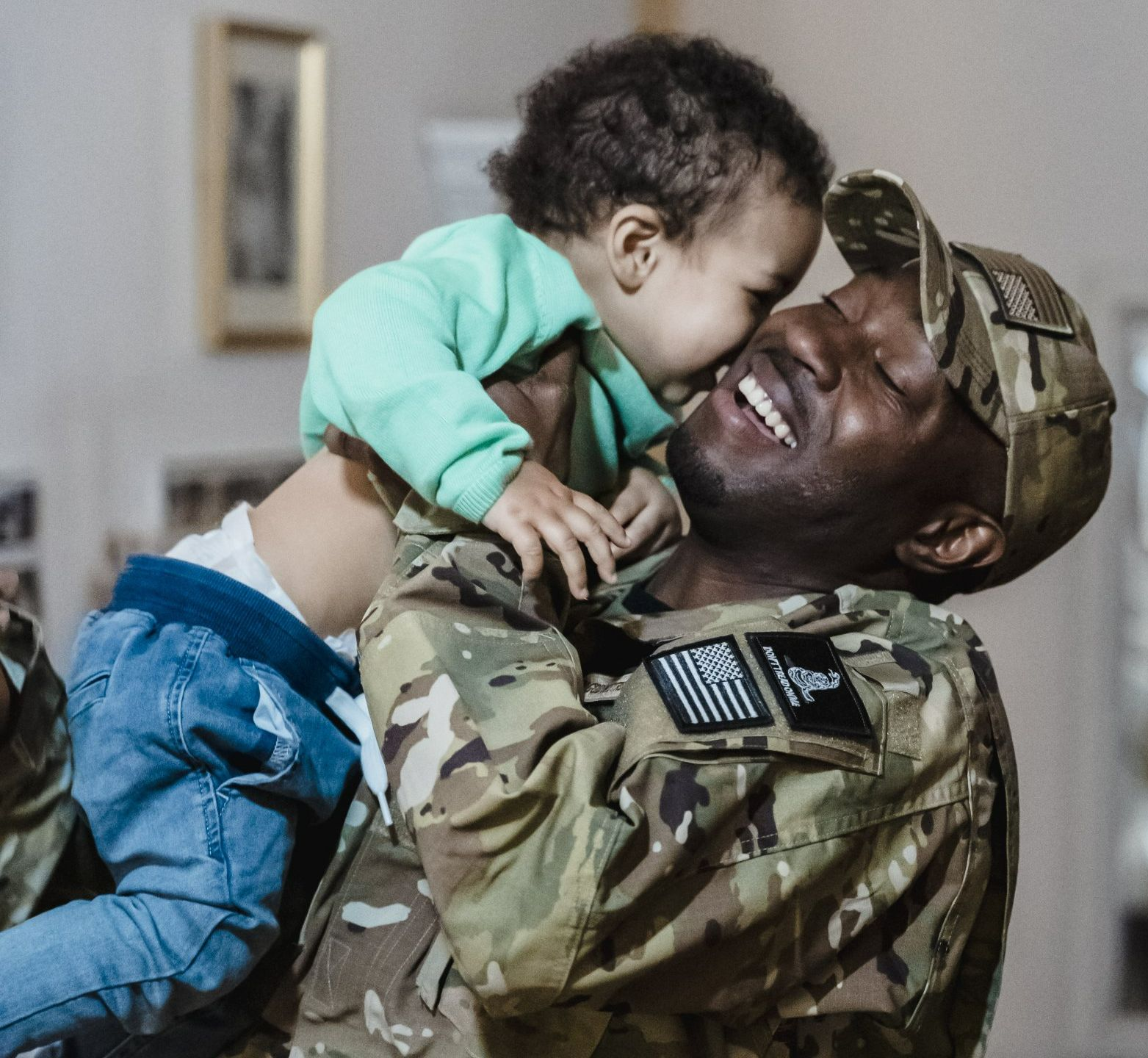 Soldier in camouflage smiling while lifting a child indoors