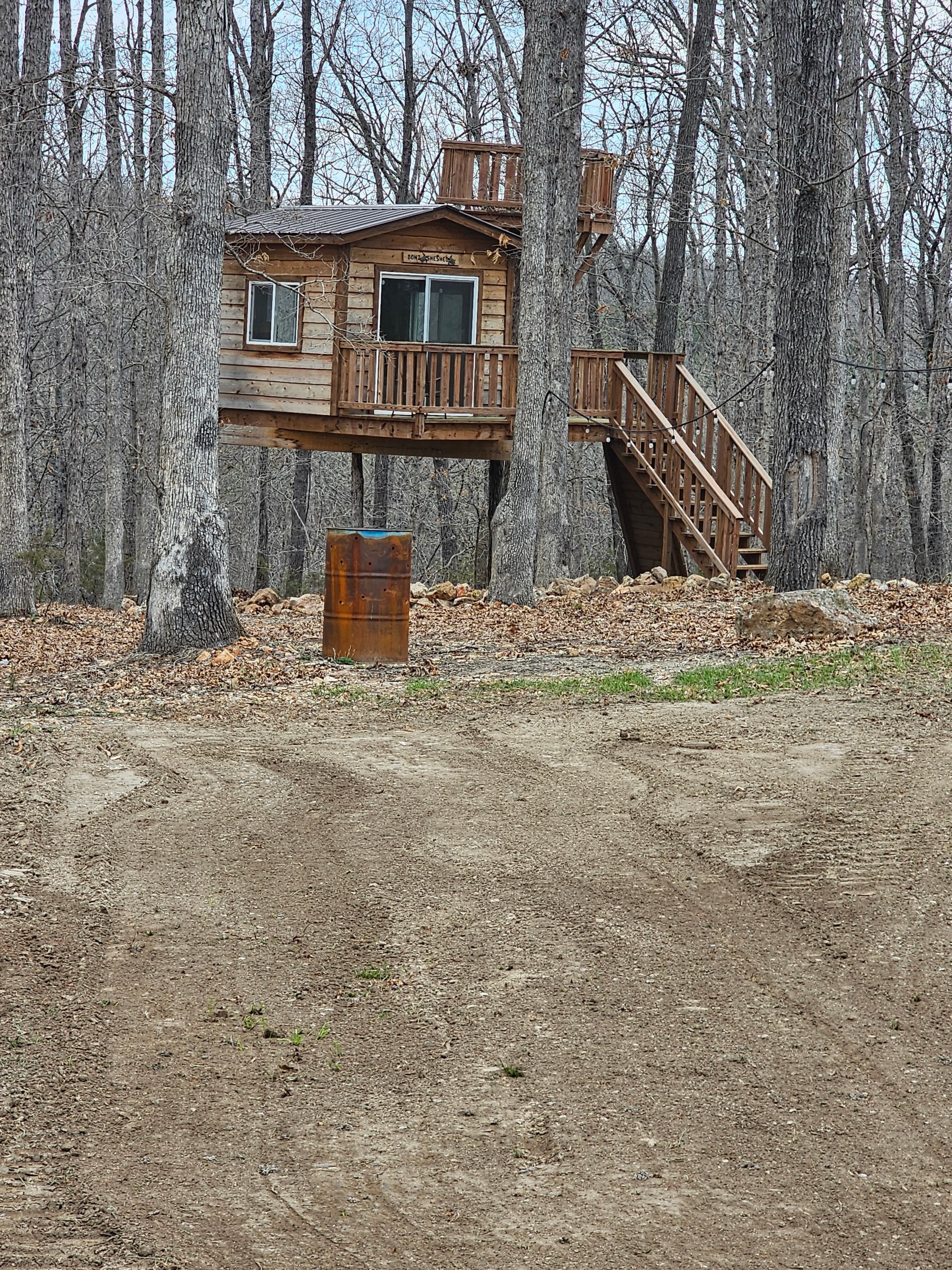 Wooden treehouse among leafless trees, with stairs and a rusty barrel in the foreground.