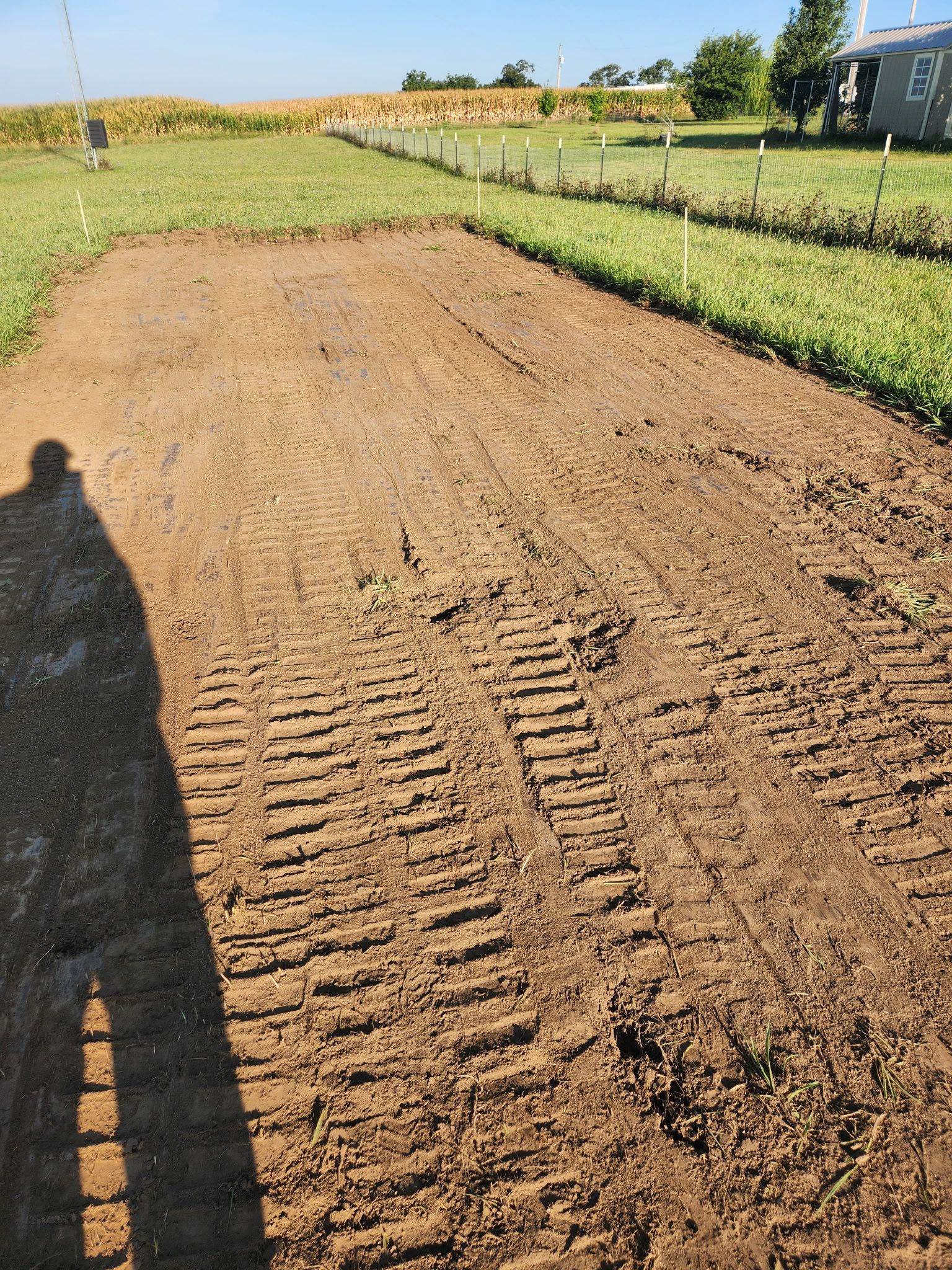 Freshly tilled brown field under bright sun, with a person’s shadow in the foreground and green crops along the edges