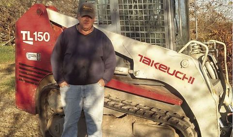 Man standing beside a red and white Takeuchi TL150 skid steer in a wooded area