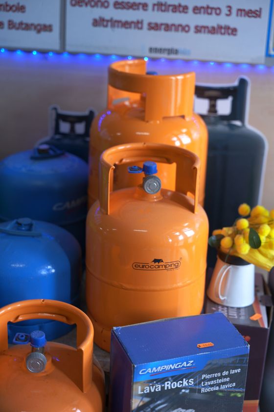 Bombole di gas GPL di colore arancione brillante impilate su uno sfondo verde, bianco e blu.