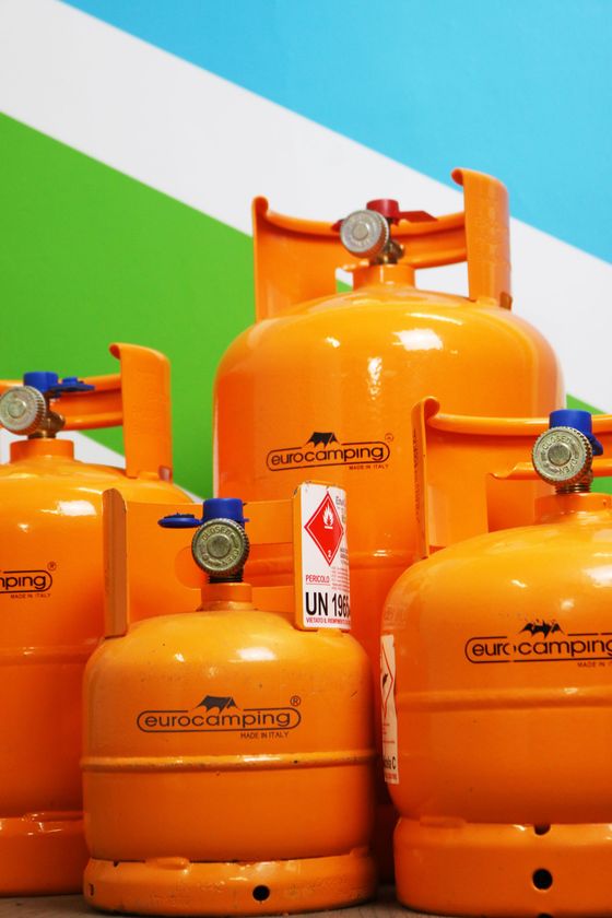 Bombole di gas GPL di colore arancione brillante impilate su uno sfondo verde, bianco e blu.