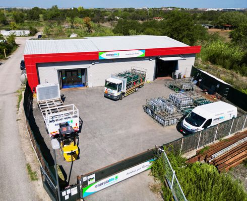 Veduta aerea di un piazzale di magazzino con camion, pallet e attrezzature industriali accanto a un edificio rosso e bianco.