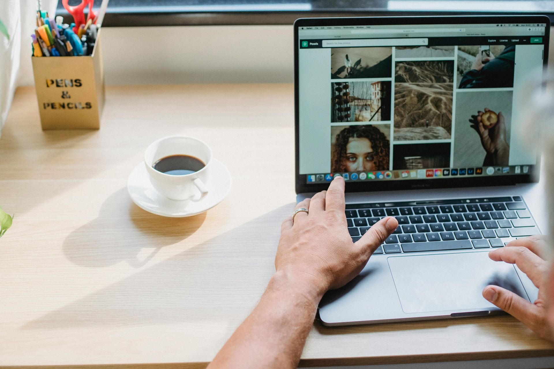 Person typing on a laptop at a desk with a coffee cup and stationery nearby, viewing a photo grid on screen
