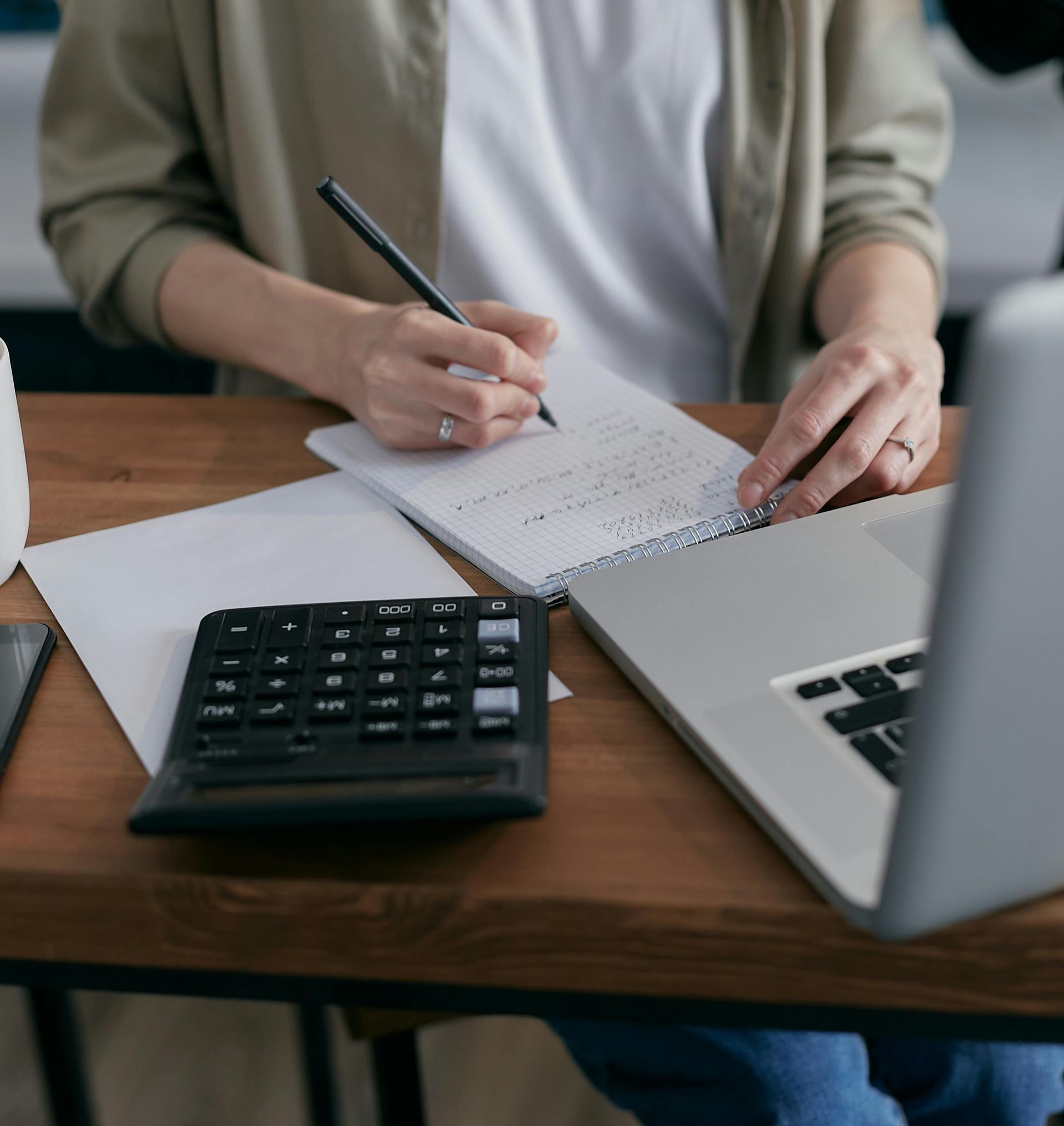 Person writing notes on a pad with calculator and laptop on a desk.