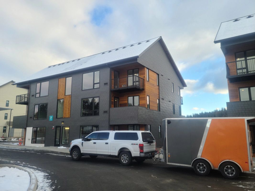 White truck towing a gray and orange trailer in front of a modern apartment building with snow-covered roof.