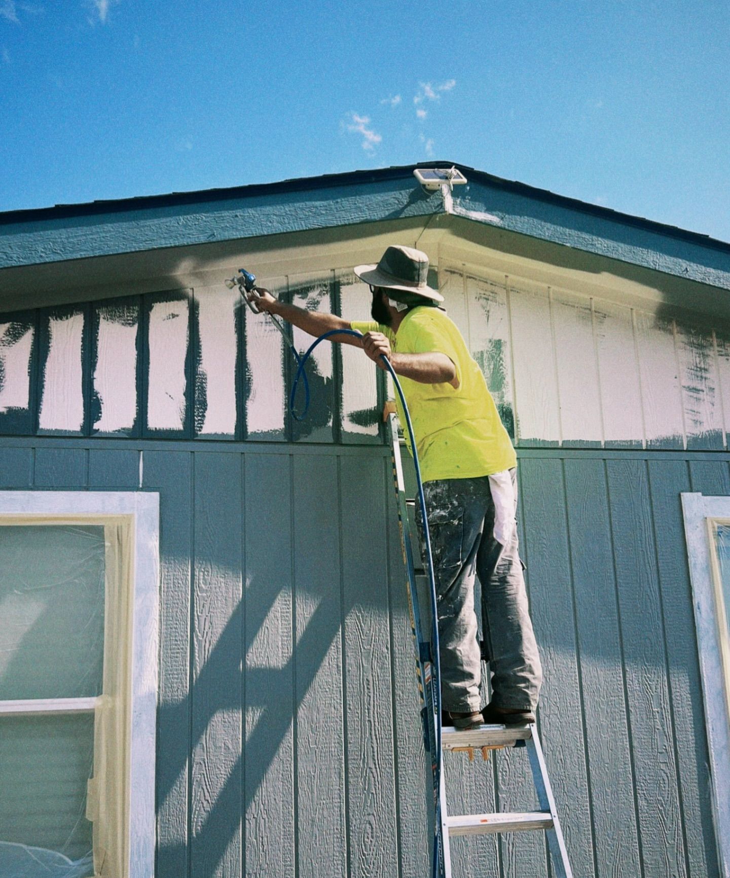 Man on ladder sprays siding of house with paint.